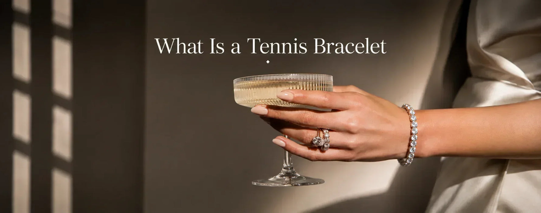 A women holding a glass of wine, wearing a tennis necklace, engagement ring and wedding band, text is what is  tennis bracelet, and in the bckgroiound there is a shadow cause by a sunrays