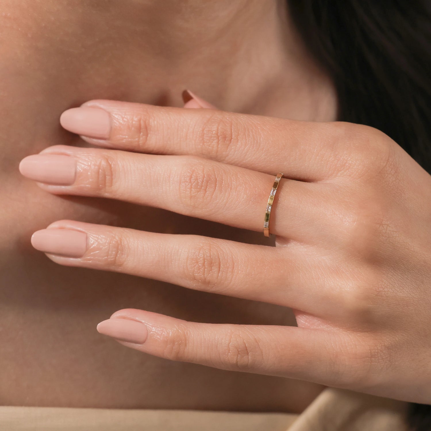Elegant thin yellow  gold eternity band with baguette diamond worn on a woman's ring finger. Soft lighting highlights the stackable ring