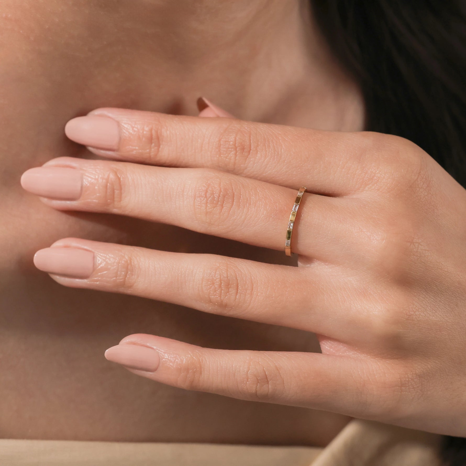 Elegant thin yellow  gold eternity band with baguette diamond worn on a woman's ring finger. Soft lighting highlights the stackable ring