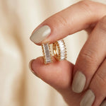 Fingers holding a pair of yellow gold huggie hoop earrings featuring a row of baguette diamonds with beaded prong settings earring.