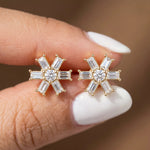 Close-up of a hand holding a pair of prong-set baguette and round diamond stud earring studs in yellow gold, arranged in a flower starburst cluster.