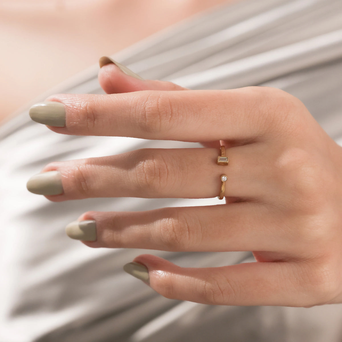 Close-up of a hand wearing a delicate gold open cuff ring with a baguette diamond and a round diamond on a soft fabric backdrop.