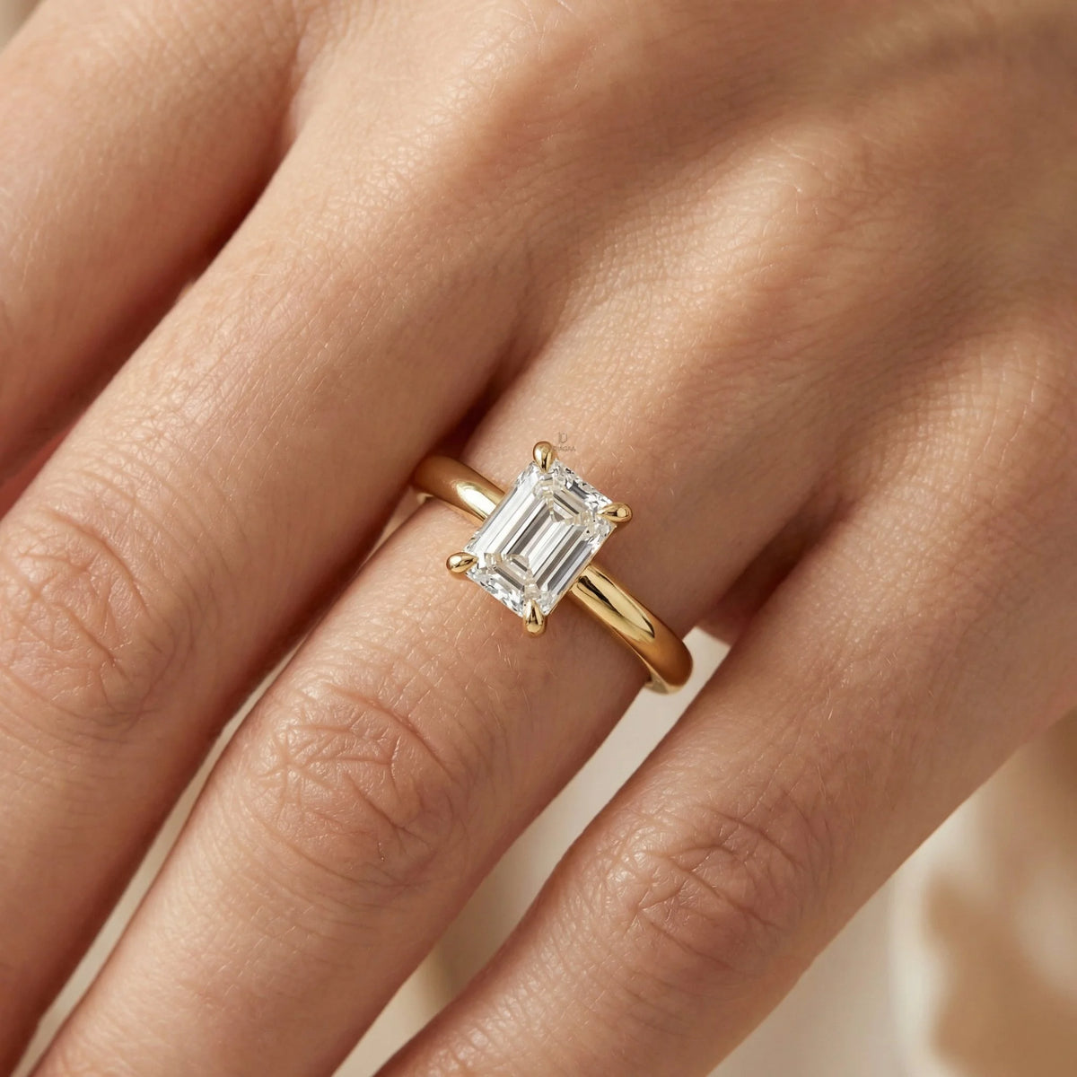 emerald cut solitaire diamond ring with claw prongs on a yellow gold band.worn in a hand.