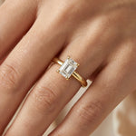 emerald cut solitaire diamond ring with claw prongs on a yellow gold band.worn in a hand.