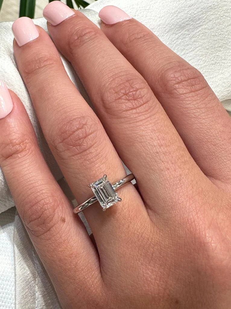 Timeless emerald-cut diamond ring on a hand with a pale pink manicure. Solitaire four-prong setting in white gold against silk fabric.