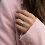 Elegant yellow gold engagement ring featuring a sparkling emerald cut diamond. Worn on a hand against a stylish pink coat lapel.