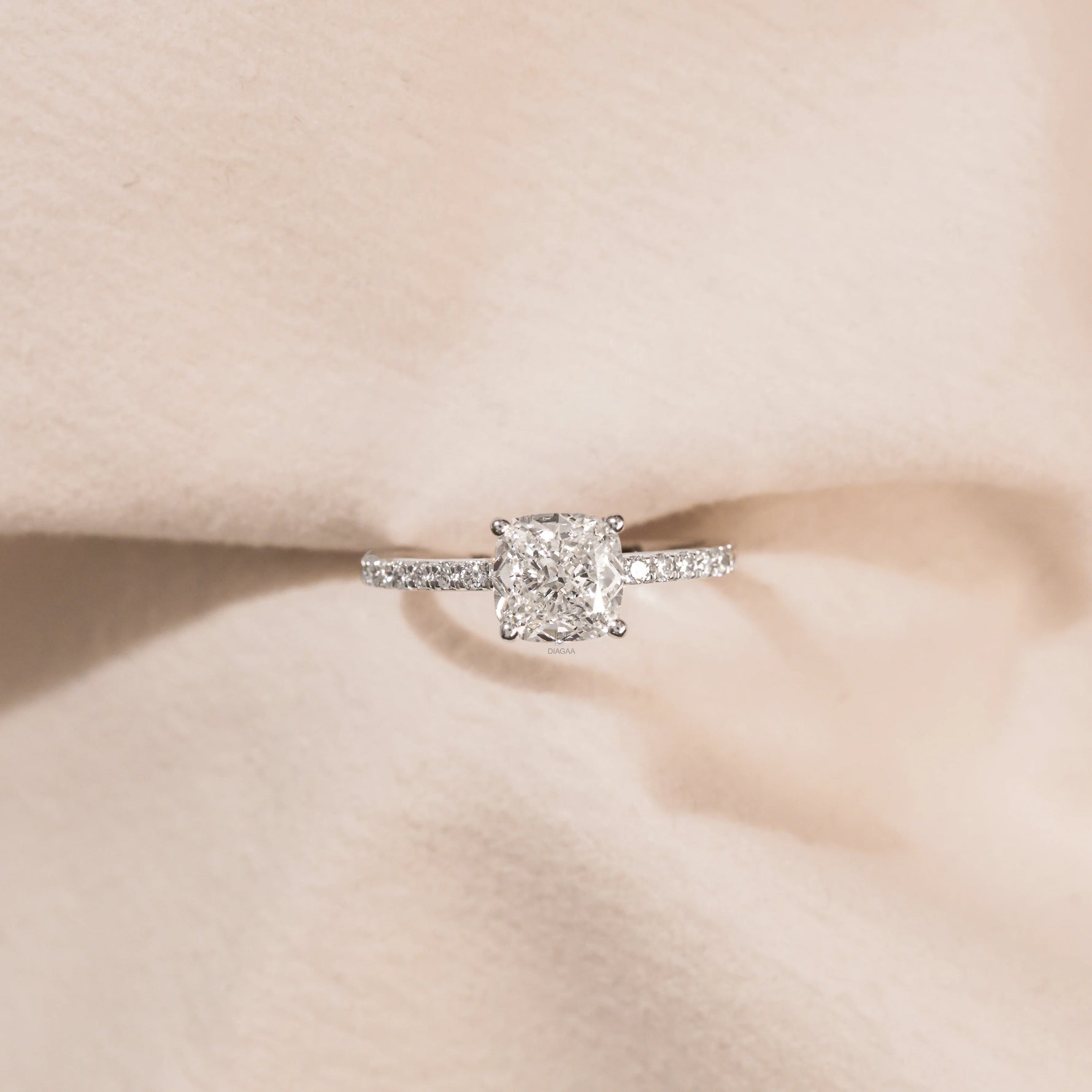 Cushion cut diamond engagement ring with a sparkling diamond pavé band resting on soft beige fabric.
