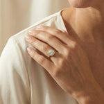 Close-up of a woman's hand wearing a yellow gold engagement ring with a round center diamond and cushion halo on beige fabric