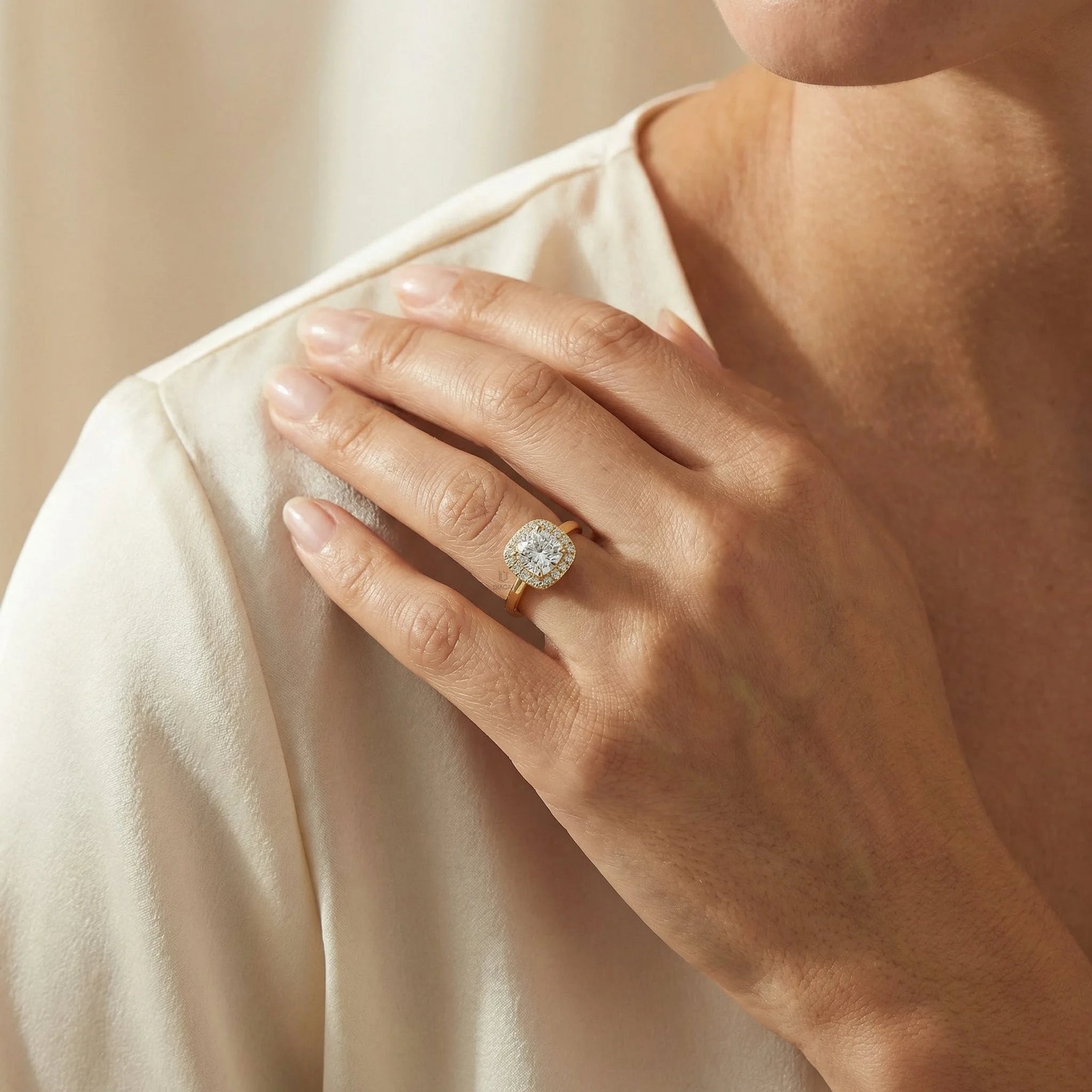 Close-up of a woman's hand wearing a yellow gold engagement ring with a round center diamond and cushion halo on beige fabric
