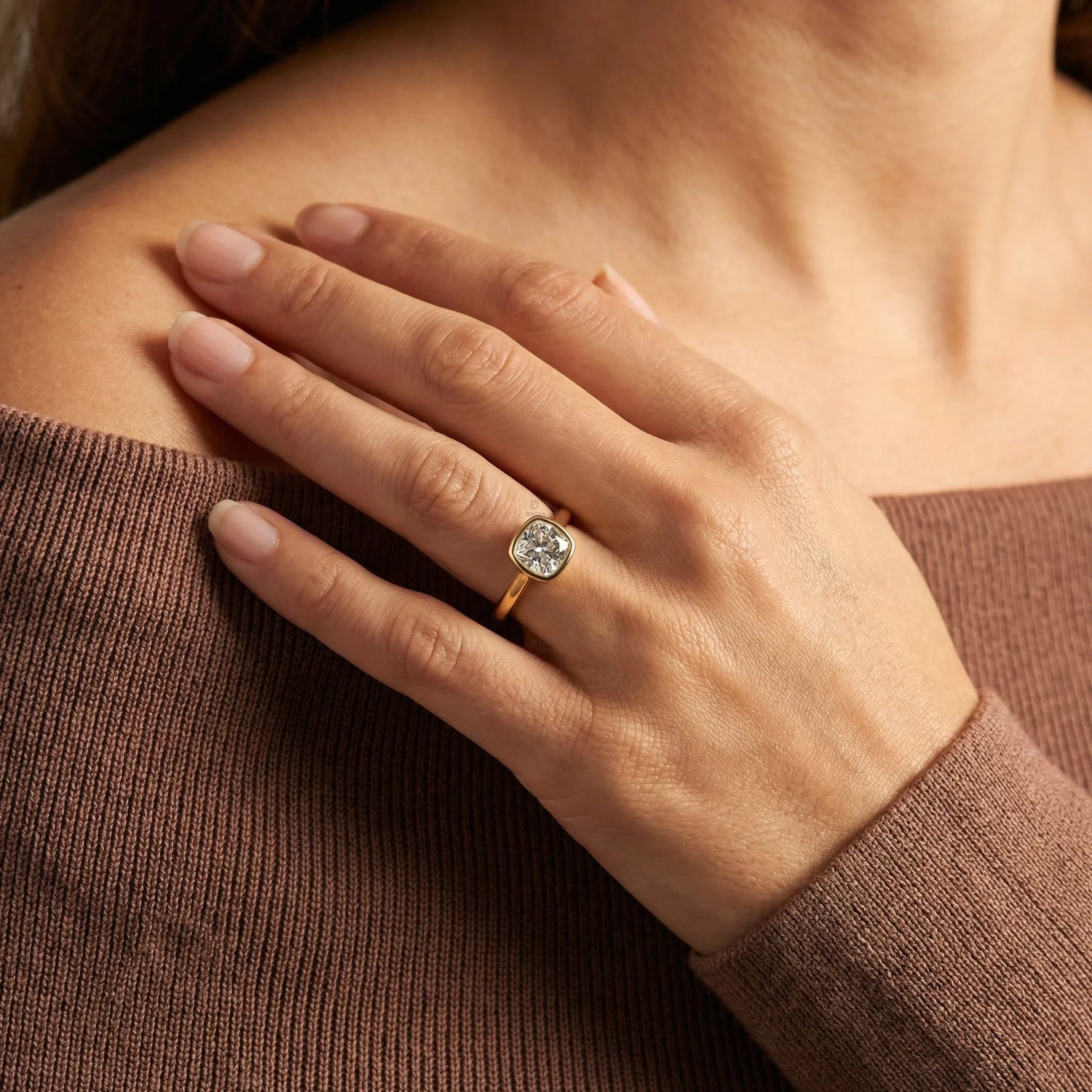 Close-up of a yellow gold cushion-cut diamond engagement ring in a bezel setting, styled on a woman's hand wearing a brown knit sweater.