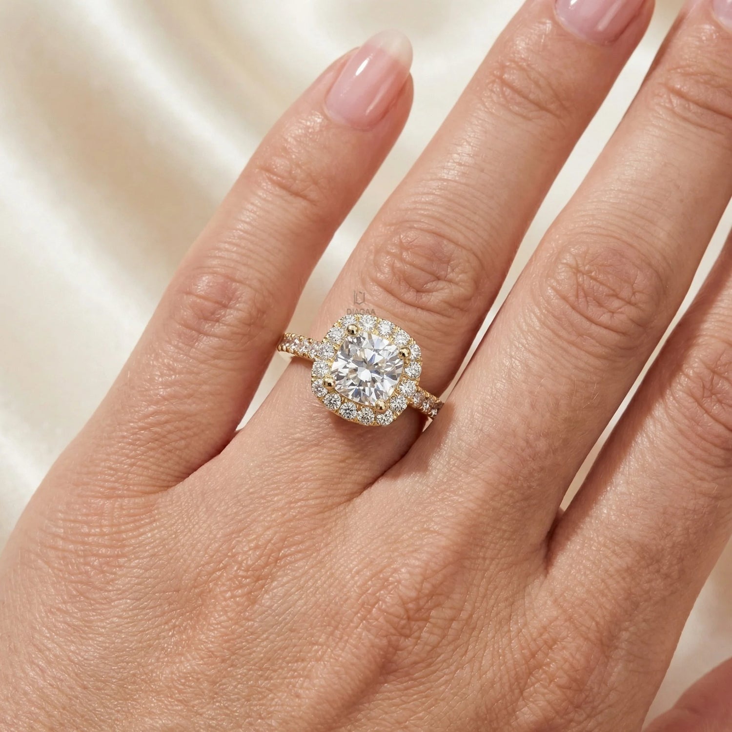 Yellow gold diamond engagement ring with a cushion-shaped halo and sparkling pave band worn on a ring finger over a cream silk backdrop.