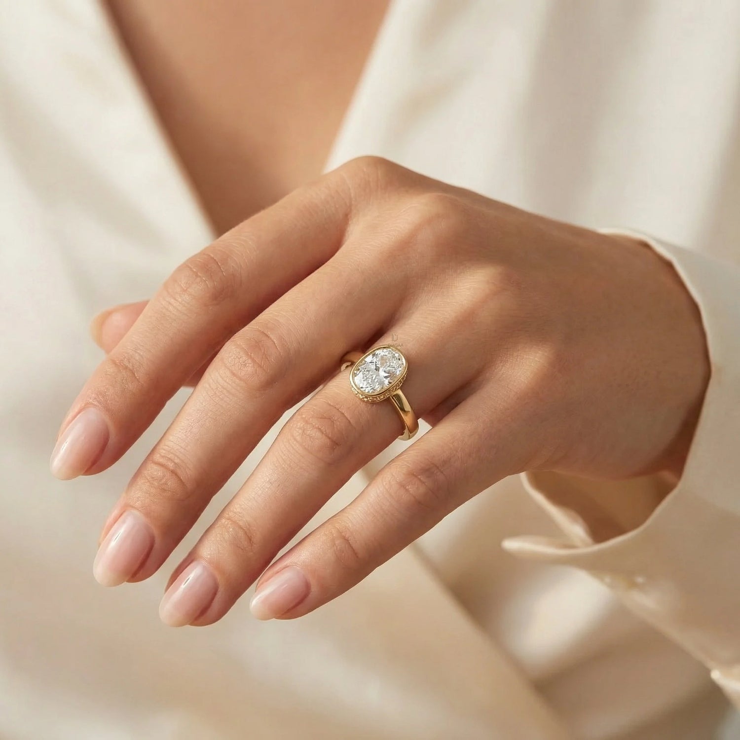 cushion cut diamond engagement ring in yellow gold with a bezel setting on a woman's hand wearing a white silk shirt.