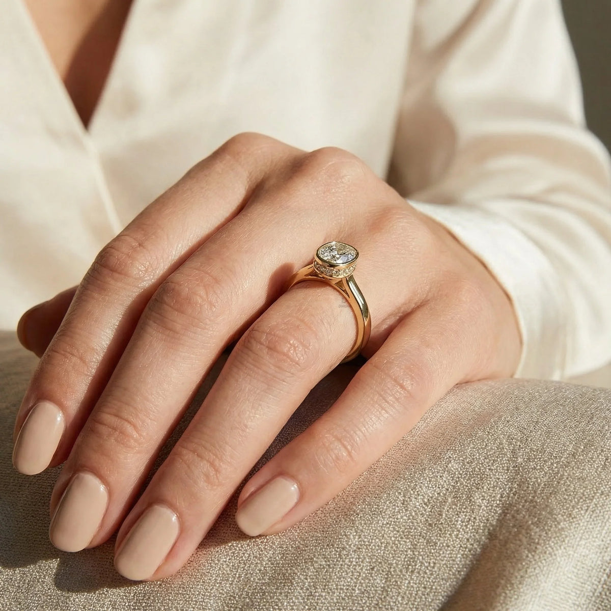 cushion cut diamond solitaire engagement ring in yellow gold with a bezel setting and hidden halo on a hand wearing a white silk shirt.