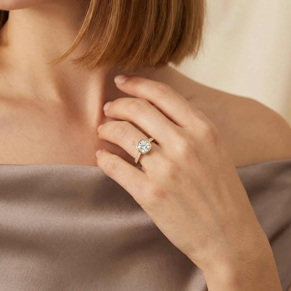 Close-up of a yellow gold cushion-cut diamond engagement ring with a modern bezel setting and sparkling pavé band, styled on a woman's hand wearing a taupe silk top.