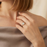 Close-up of a yellow gold cushion-cut diamond engagement ring with a modern bezel setting and sparkling pavé band, styled on a woman's hand wearing a taupe silk top.