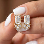 Pair of emerald and round diamond stud earring designs held in fingers, crafted in yellow gold with a delicate prong setting.