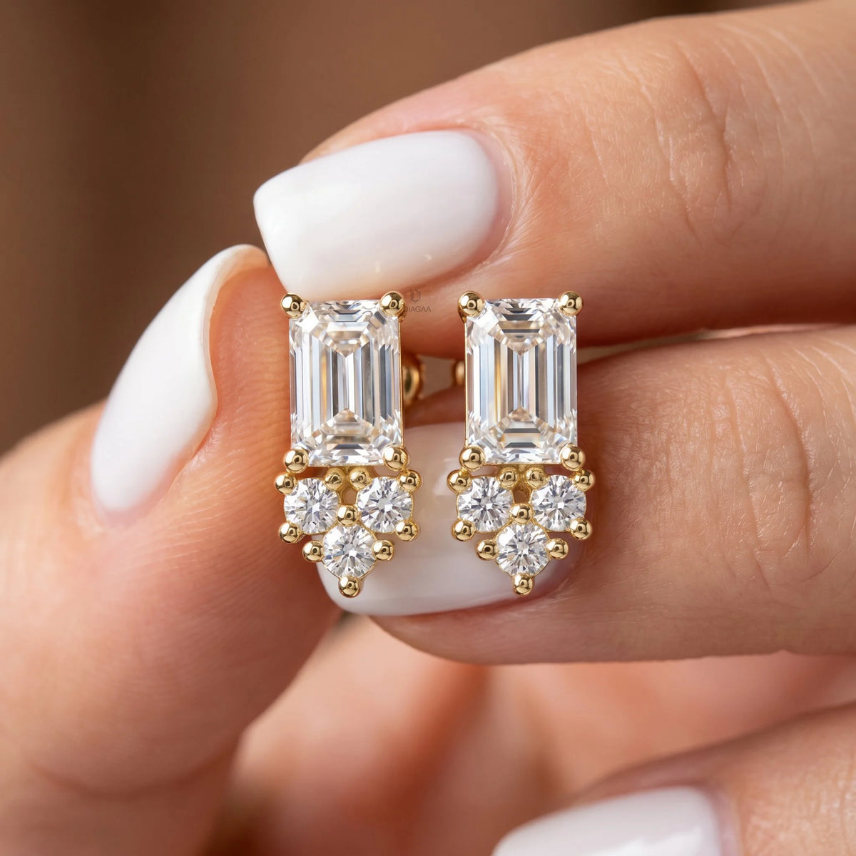 Pair of emerald and round diamond stud earring designs held in fingers, crafted in yellow gold with a delicate prong setting.