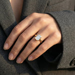Close-up of a hand wearing a stunning emerald-cut diamond engagement ring in a yellow gold bezel setting, resting against a grey blazer.