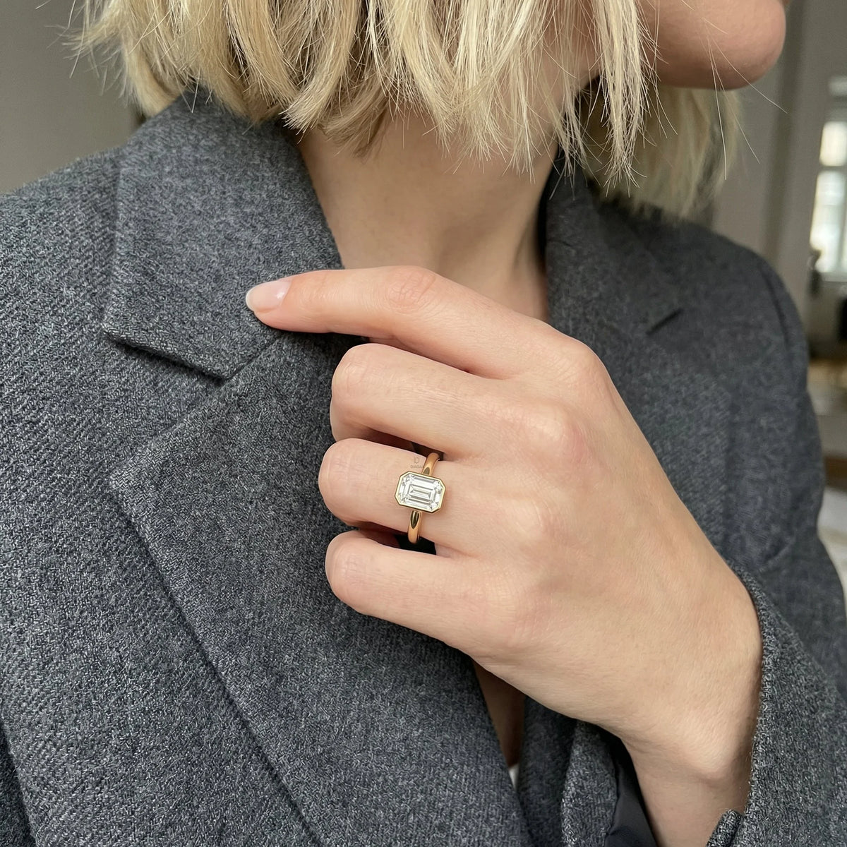 Minimalist yellow gold bezel solitaire ring featuring a rectangular emerald diamond, displayed gracefully on a woman’s ring finger for a modern bridal look.