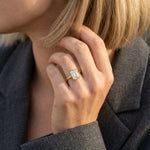 Woman wearing a yellow gold bezel set emerald-cut diamond engagement ring on her ring finger, posing in a dark grey suit jacket in natural sunlight.