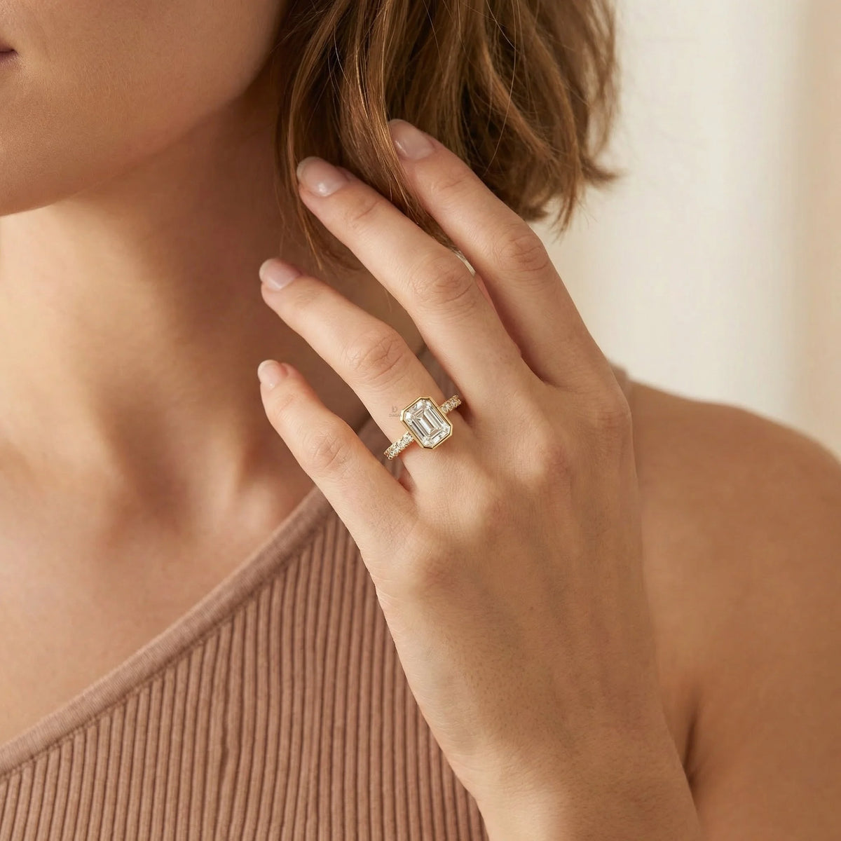 A luxury yellow gold emerald diamond bezel set engagement ring featuring a prominent round cut side stone pave set band frame, modeled elegantly on a hand.