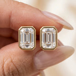 A hand holding a pair of emerald cut diamond stud earrings in a polished yellow gold bezel setting earring.