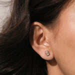  Woman running a hand through dark brown hair to show a yellow gold bezel set emerald cut diamond stud earring on her ear lobe.