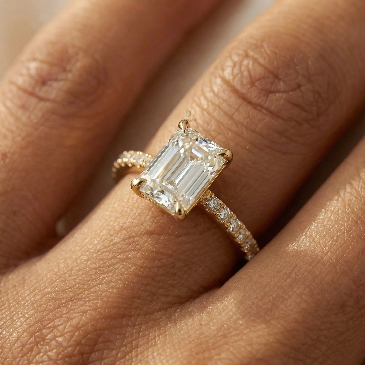 Yellow gold engagement ring featuring an emerald cut diamond and round cut side stone pave band, modeled on a hand over white satin