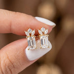 Yellow gold emerald, marquise and round diamond stud earring pair held between fingers, featuring a secure prong setting.