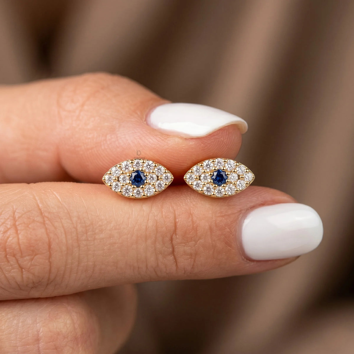 Pair of gold-plated evil eye stud earrings with pavé diamond accents and a central blue sapphire stone, held between fingers with white manicured nails.
