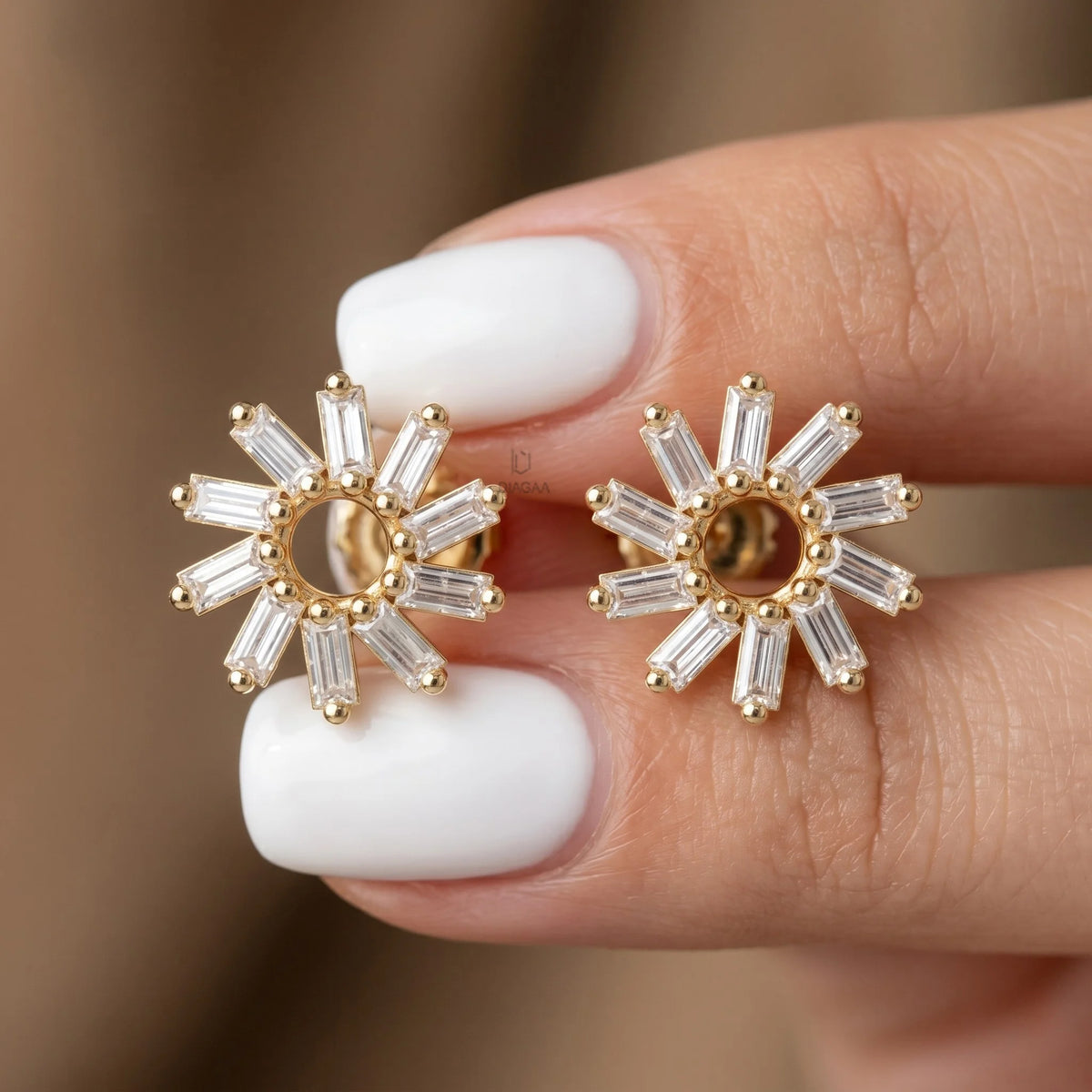 A hand with crisp white manicured nails holding a pair of yellow gold sunburst stud earrings encrusted with radiating baguette diamonds around an open circle.