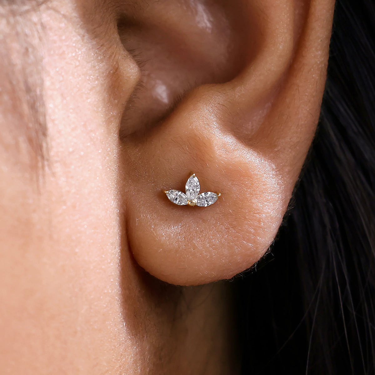 A close-up view of a woman wearing a delicate marquise diamond stud earring set in solid 14K yellow gold, showcasing a three-stone floral cluster design that is securely prong-set on the earlobe.