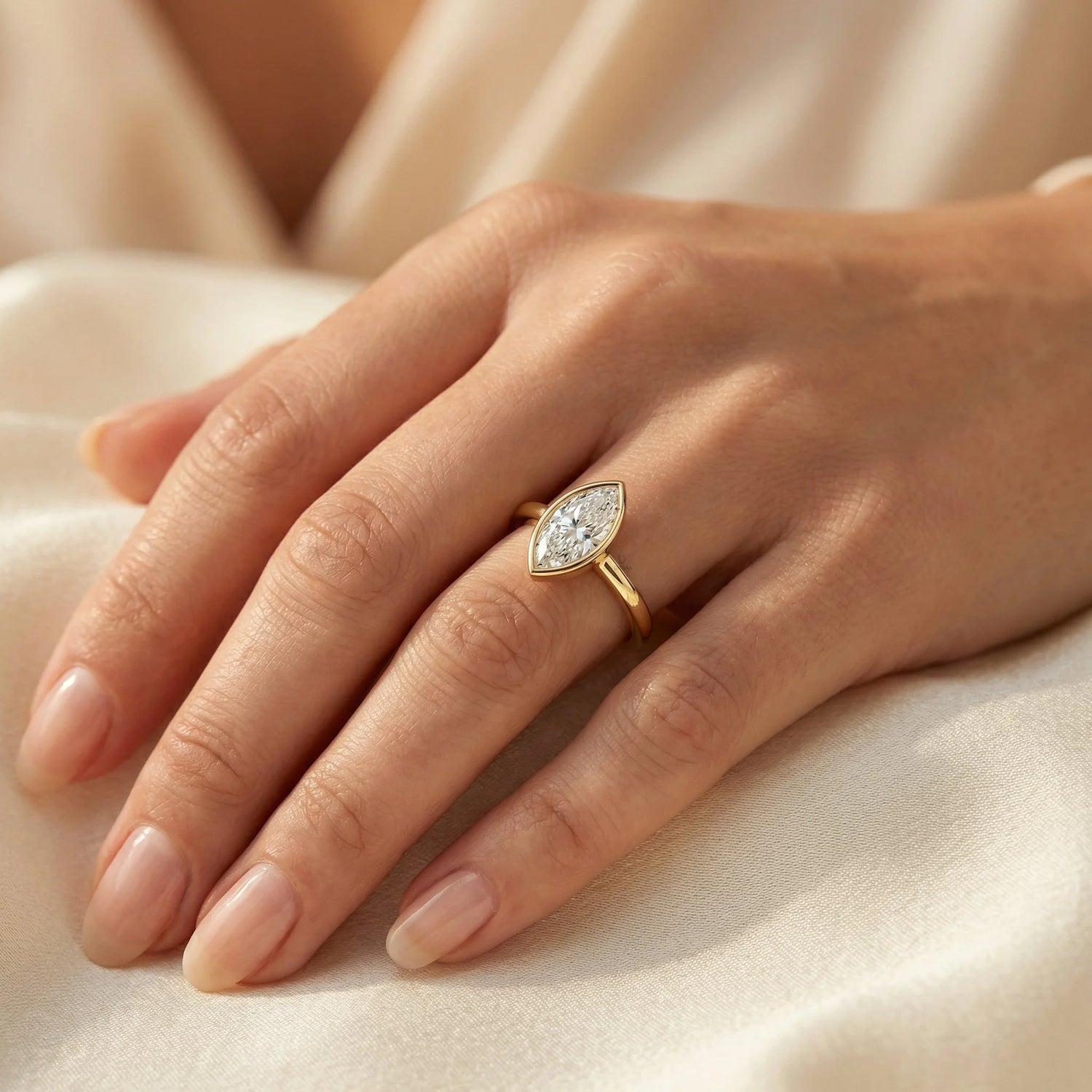 Close-up of a yellow gold marquise-cut diamond engagement ring in a bezel setting, styled on a woman's hand resting on a soft cream fabric background.