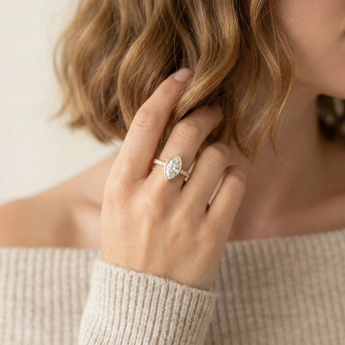 Close-up of a 14k yellow gold marquise-cut diamond engagement ring with a modern bezel setting and shimmering pavé band, styled on a woman's hand wearing a beige knit sweater.