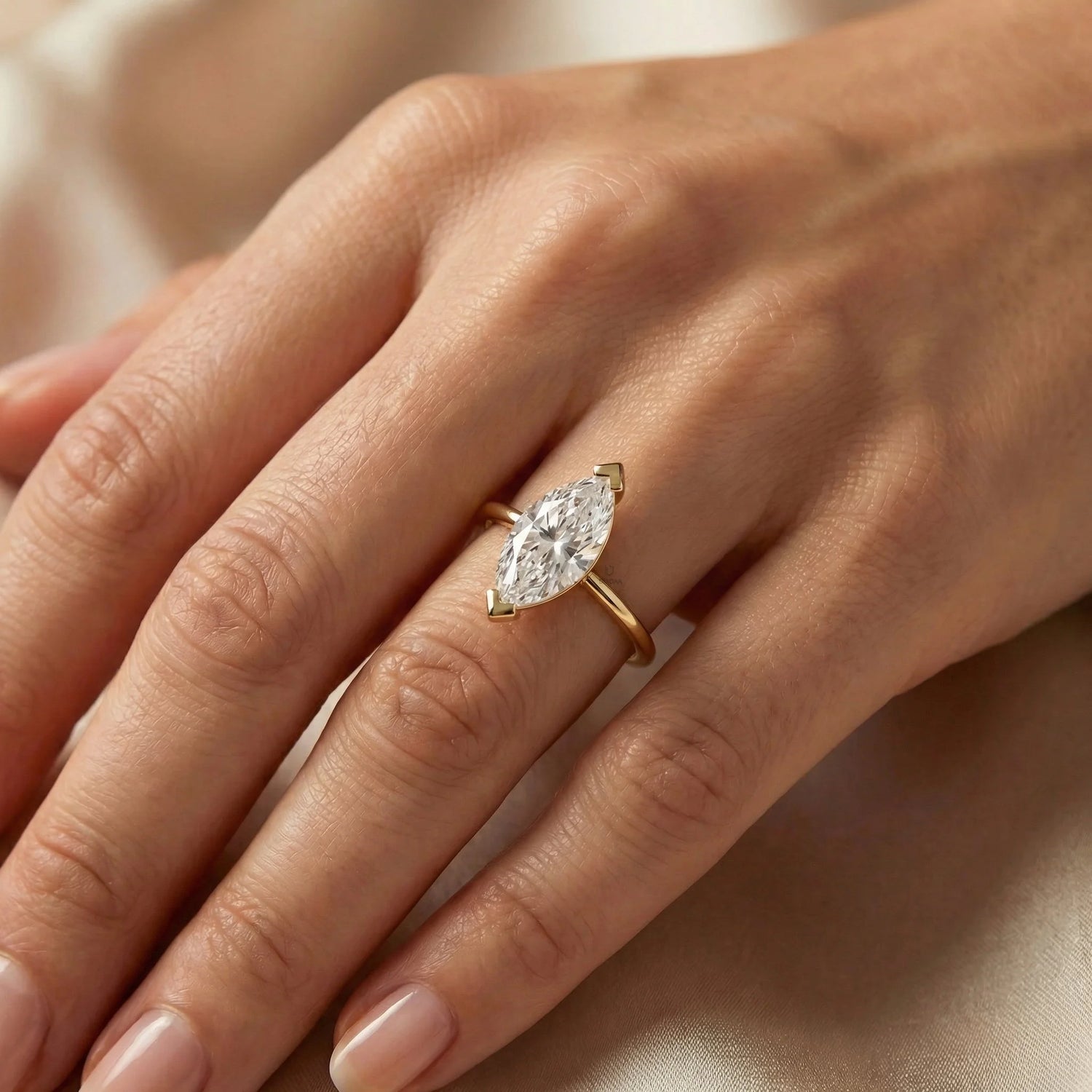 Solitaire marquise diamond engagement ring in a yellow gold v-prong setting, worn on a ring finger against a neutral satin backdrop.