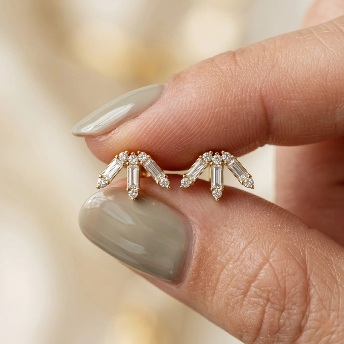 Close-up shot of a hand holding a pair of prong-set baguette and round diamond stud earring studs in yellow gold, showcasing a geometric arch design.