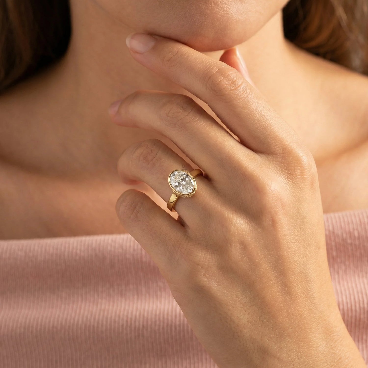 Close-up of a woman wearing an oval-cut diamond engagement ring in a yellow gold bezel setting, styled with a soft pink top.