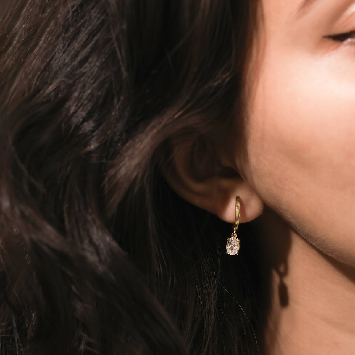 A woman's profile highlighting a dainty yellow gold drop earring with an oval-cut diamond hanging from a huggie hoop dangling earring 