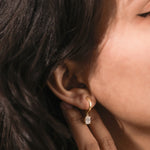 A woman's profile highlighting a dainty yellow gold drop earring with an oval-cut diamond hanging from a huggie hoop earring .