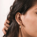Close-up model shot showcasing a yellow gold huggie hoop earring with a dangling prong-set oval diamond charm on an ear lobe.