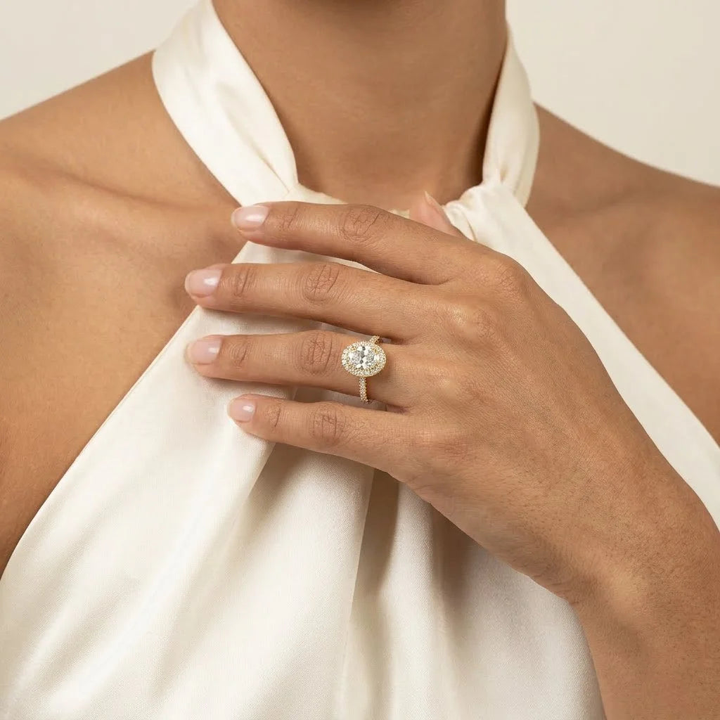 Close-up of a hand wearing a stunning yellow gold halo engagement ring with a brilliant center diamond, paired with an elegant white satin halter top.