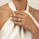 Close-up of a hand wearing a stunning yellow gold halo engagement ring with a brilliant center diamond, paired with an elegant white satin halter top.
