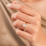 Woman's hand wearing a solitaire oval-cut diamond engagement ring in yellow gold, set against beige silk backdrop.