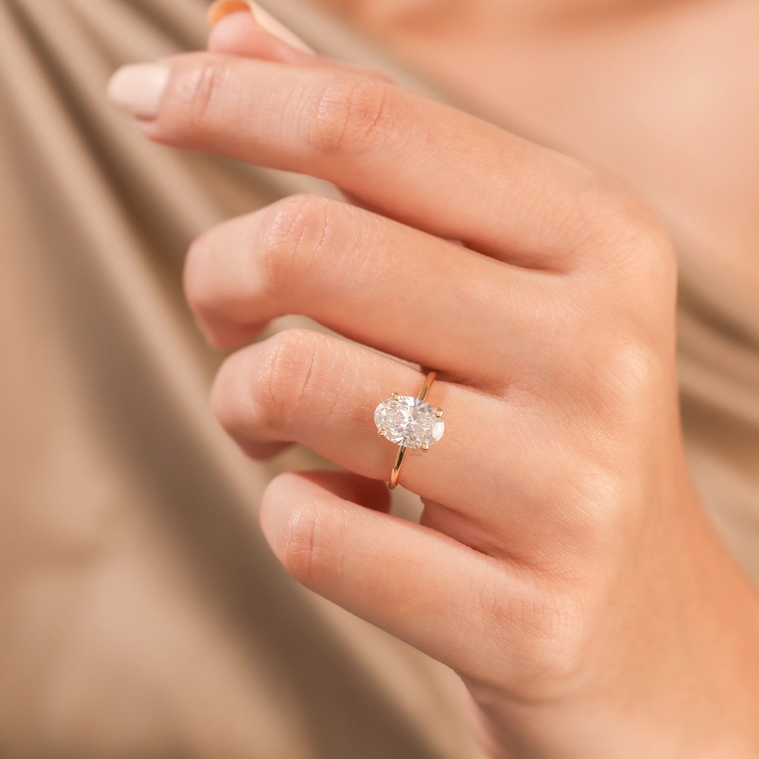 Woman's hand wearing a solitaire oval-cut diamond engagement ring in yellow gold, set against beige silk backdrop.