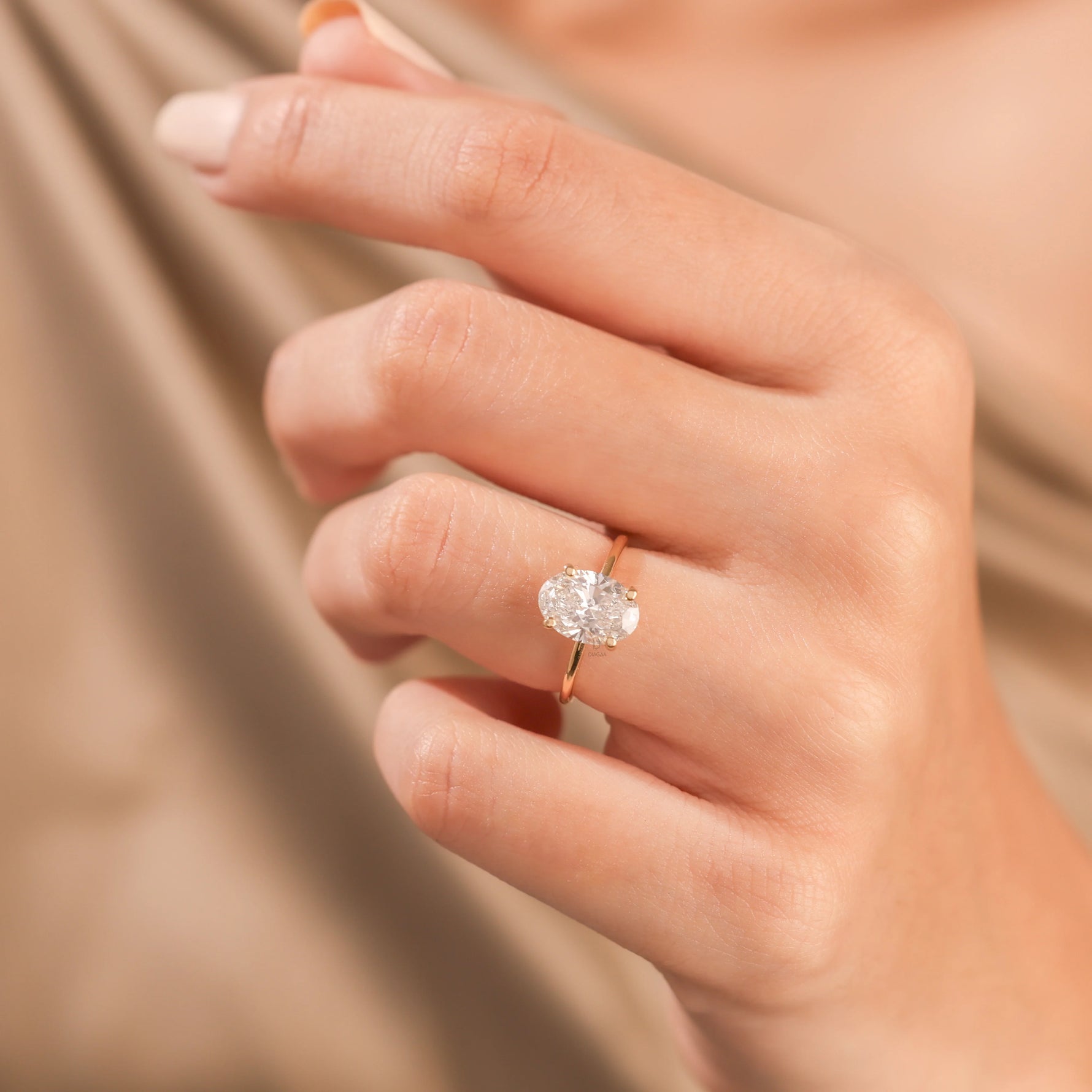 Woman's hand wearing a solitaire oval-cut diamond engagement ring in yellow gold, set against beige silk backdrop.