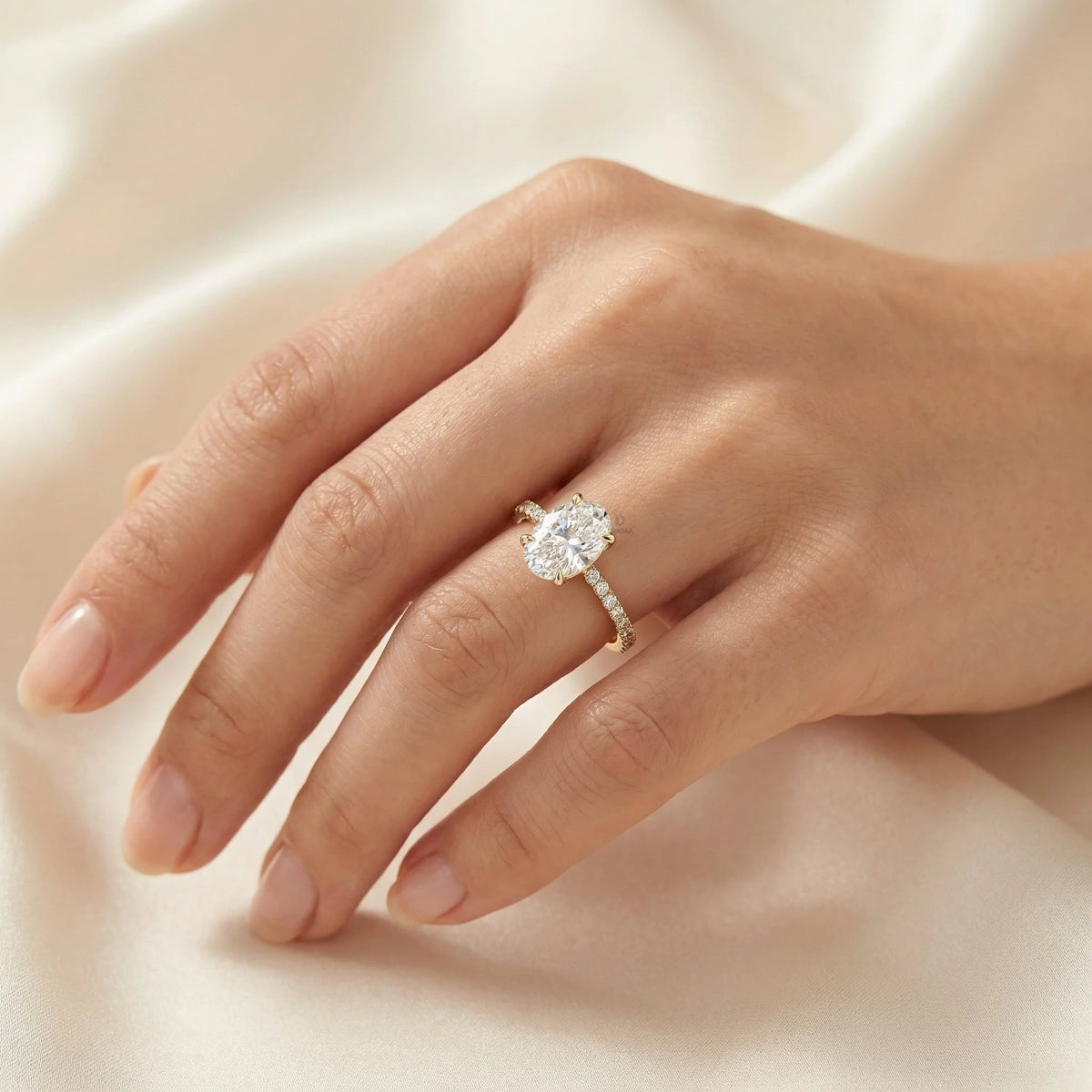 Stunning oval-cut diamond engagement ring with a pavé yellow gold band worn on a hand against a silk background.