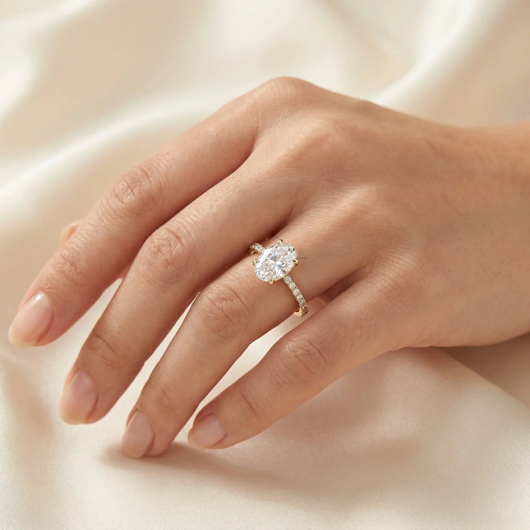 Stunning oval-cut diamond engagement ring with a pavé yellow gold band worn on a hand against a silk background.