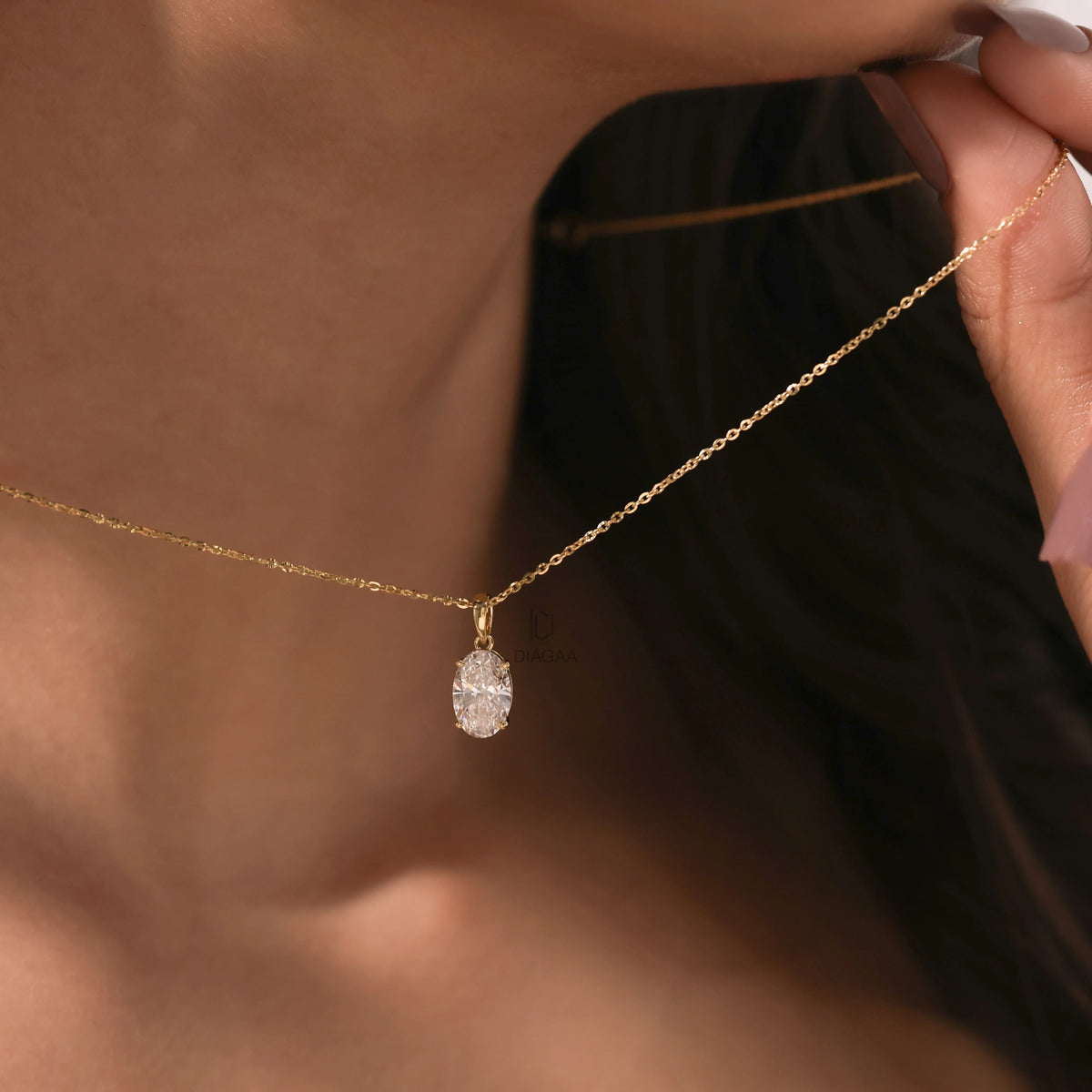 Woman gently holding the fine chain of a delicate yellow gold Oval diamond necklace, featuring a brilliant prong-set pendant.