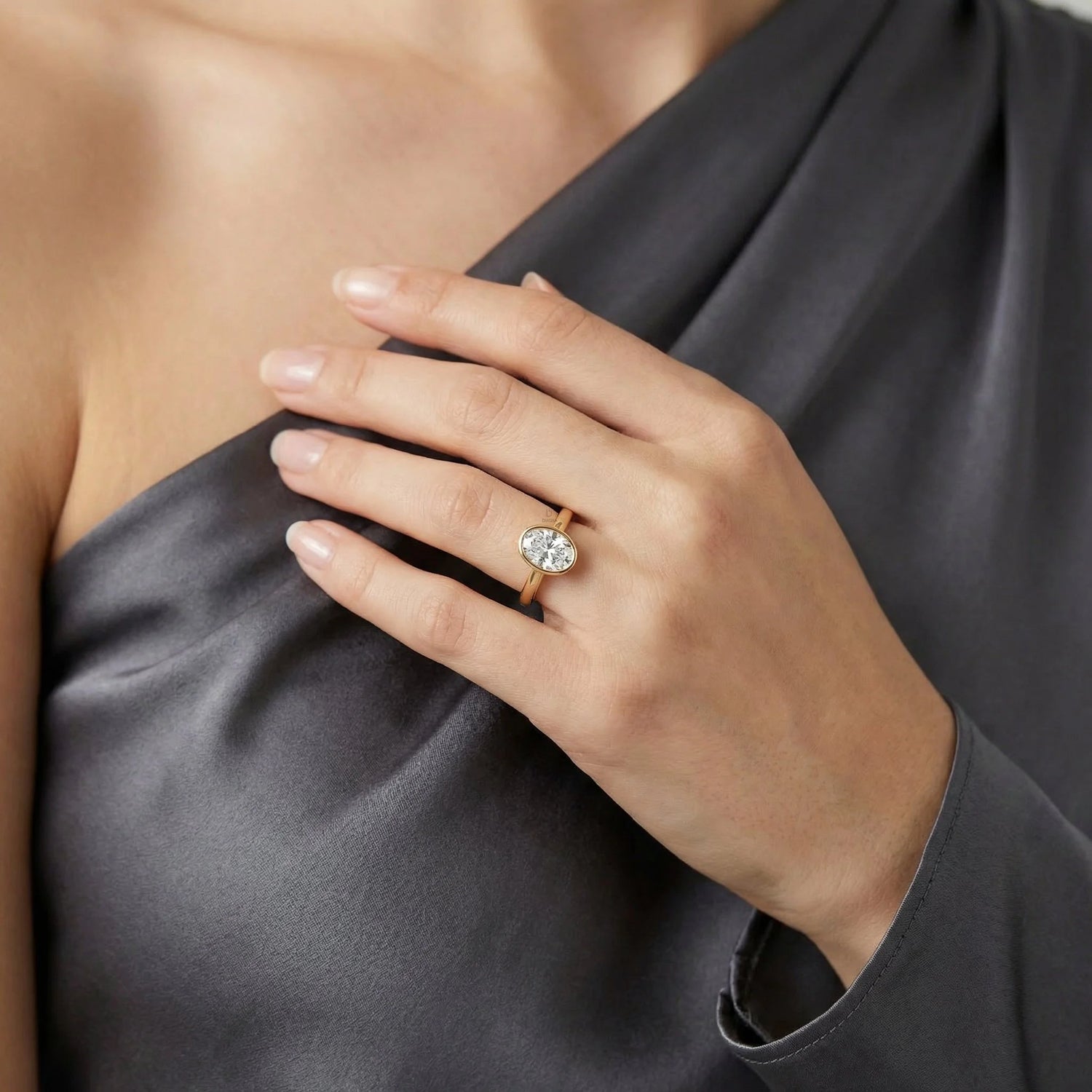 Close-up of a yellow gold oval-cut diamond engagement ring in a bezel setting, styled on a woman's hand wearing a dark grey satin sleeveless top.