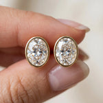 Close-up of fingers holding a pair of large yellow gold bezel-set oval diamond stud earrings to show scale and brilliance earring.